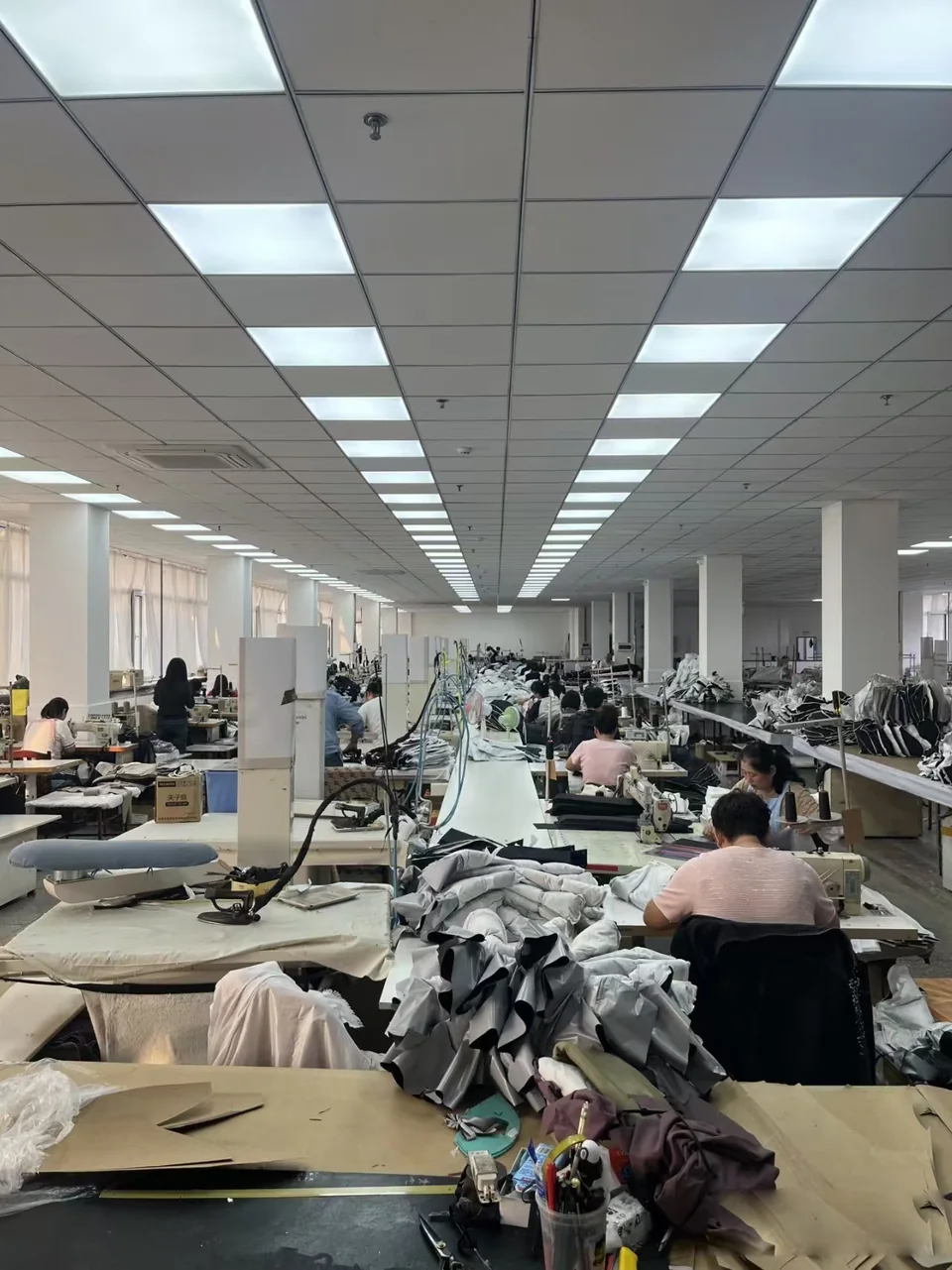 A long view along sewing tables with workers, fabric pieces and overhead lights